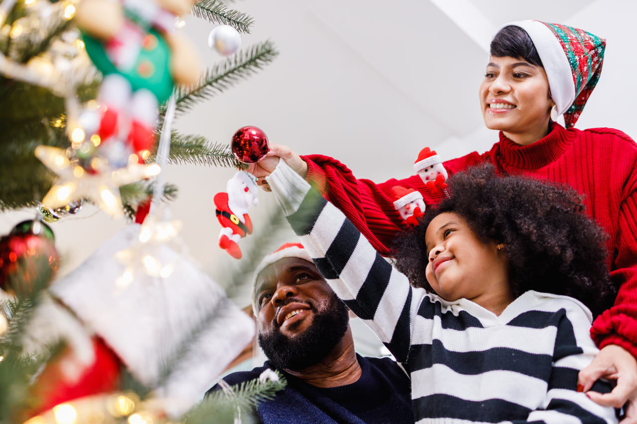 Família decorando uma árvore de Natal, com um adulto e uma criança pendurando enfeites enquanto sorriem.