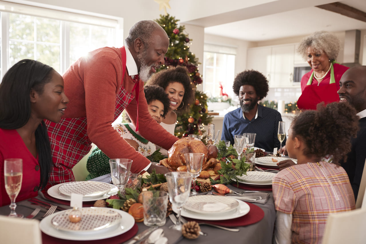 Família reunida em volta de uma mesa de Natal, com o avô colocando um peru assado no centro. Todos sorriem enquanto se preparam para a refeição, com uma árvore de Natal decorada ao fundo.