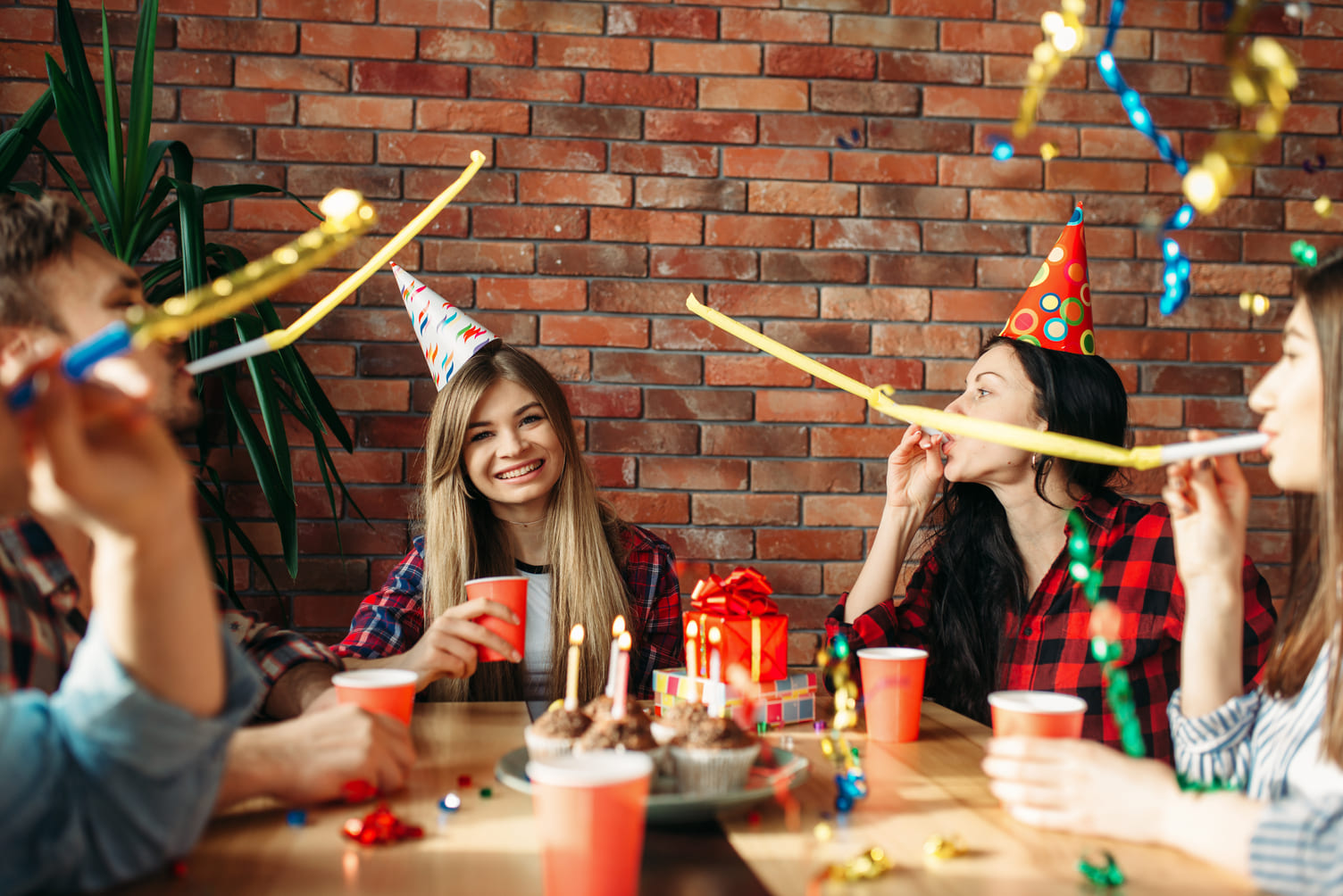 grupo de jovens está celebrando em uma festa, com chapéus coloridos de festa, bebidas em copos vermelhos e bolinhos decorados com velas acesas. Todos estão sorrindo e se divertindo, enquanto tocam em instrumentos de festa. A imagem captura um momento de alegria em um ambiente descontraído, com uma parede de tijolos ao fundo, decorada com serpentinas e balões.